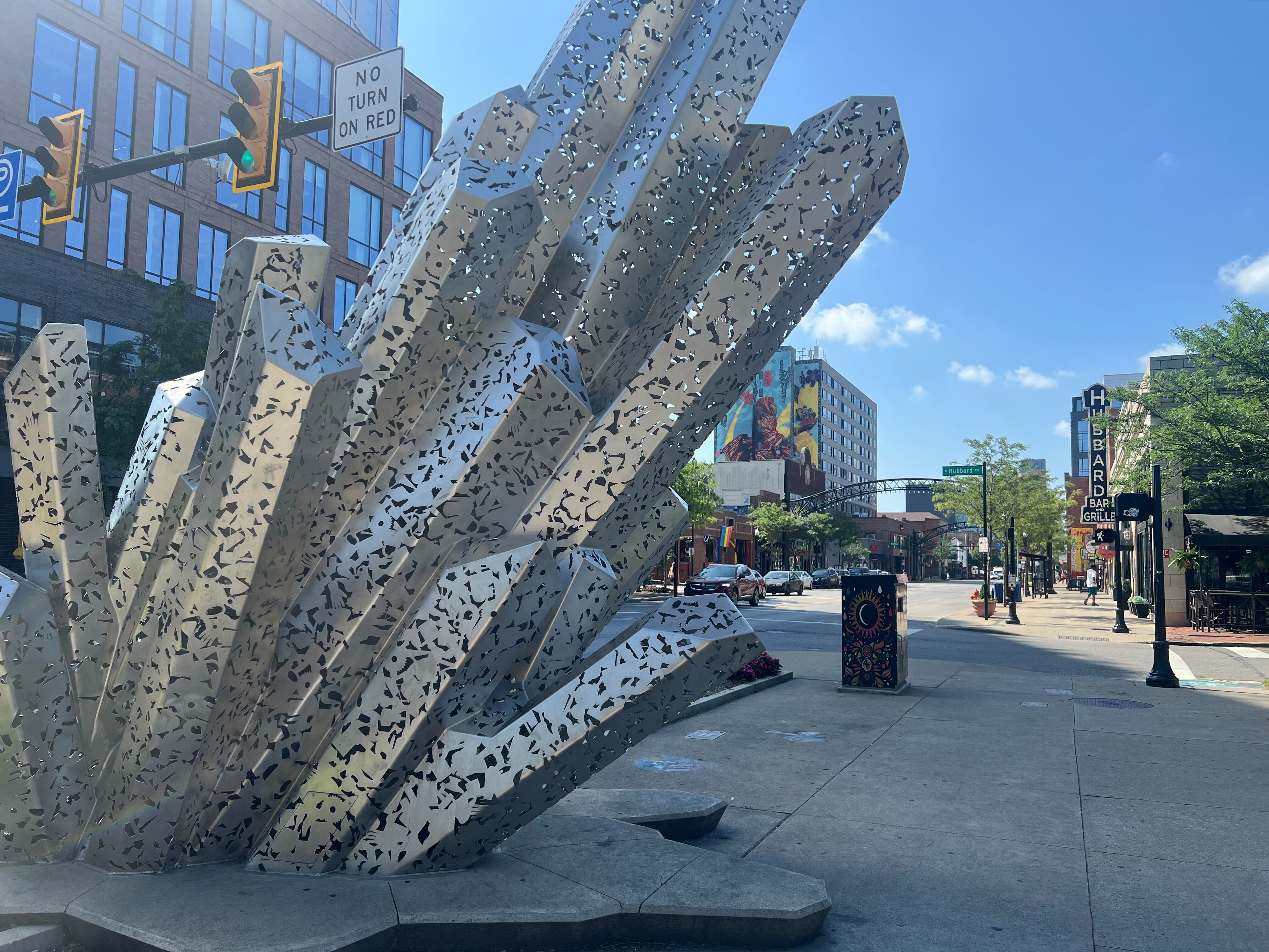 A photograph of an art piece on High St in Columbus. The piece is a sculpture of a crystal structure made of polished silver metal with patterned holes cut from it.